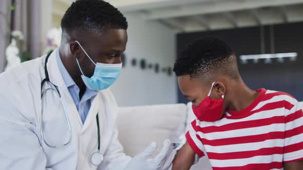 African american doctor wearing face mask injecting covid-19 vaccine into a boy at home