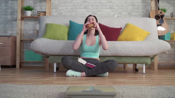 Young Sporty Woman Sitting on the Floor Looking at the Bathroom Scales Eating a Burger