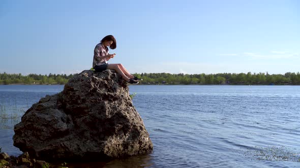 A Young Woman Sits on a Stone By the River with a Phone in Her Hands. The Girl Is Texting in the