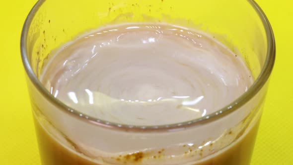 Detail shot of spoon creating a coffee whirlpool in a transparent glass, yellow background. Macro sh