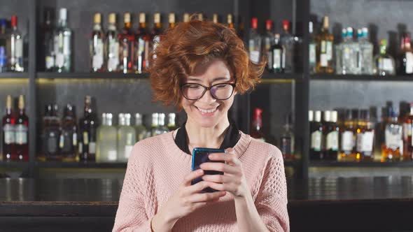 Red-haired Woman Using Smartphone in Restaurant Standing Near Bar Stand