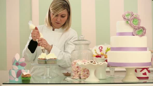 Young Pastry Chef with Pastry Bag is Garnishing Cup Cakes with Cream and Chocolate Flakes in Her