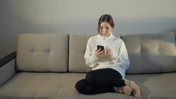 Young Woman in Sweater Sitting on Sofa Checking Her Cell Phone