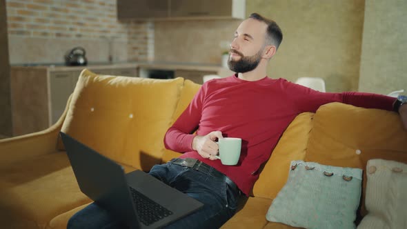 Bearded Man Having Coffee or Tea Pause While Using Laptop While at Home