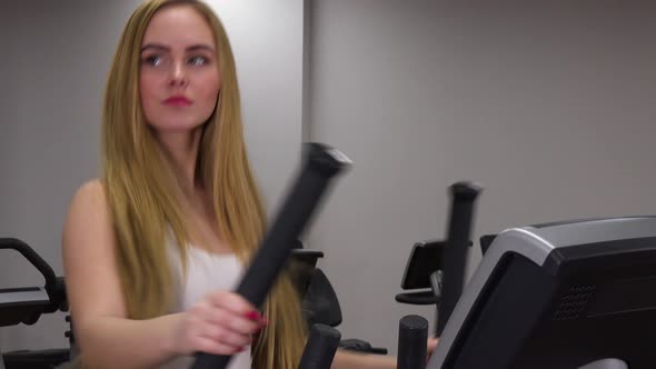 A Young Beautiful Woman Trains on an Elliptical Trainer in a Gym and Smiles at the Camera - Closeup