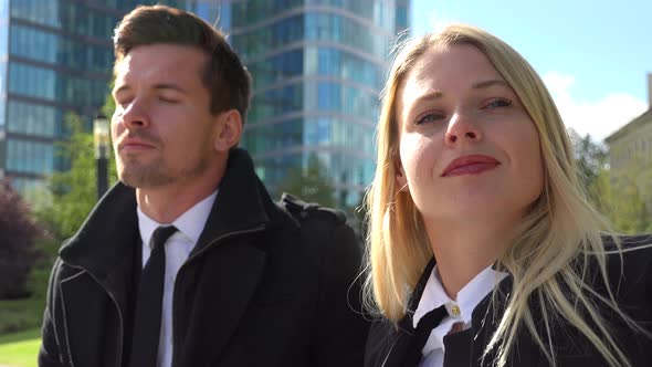 A Businessman and a Businesswoman Sit on a Bench in an Urban Area and Enjoy the Sun - Closeup