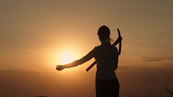 Silhouette Businesswoman in Helmet with Pickaxe on Shoulder Stands on Hill Background Sunset and