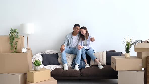 Joyful Amazed Happy Multiracial Young Couple in Love Sitting on Sofa in Living Room of Their New