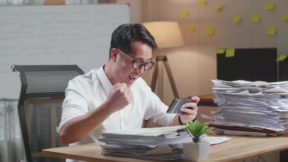 Happy Asian Man Celebrating Winning Game On Smartphone After Working With Documents At The Office