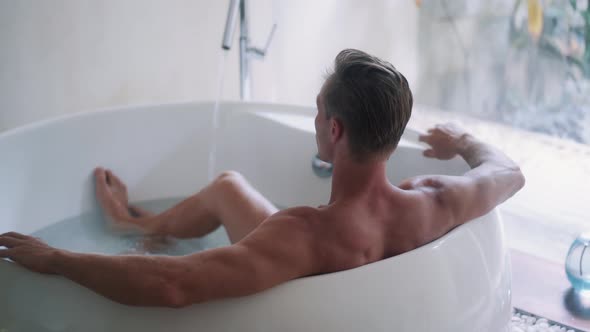 Sporty Guy with Muscular Body Rests in Large Bathtub at Home Backside View