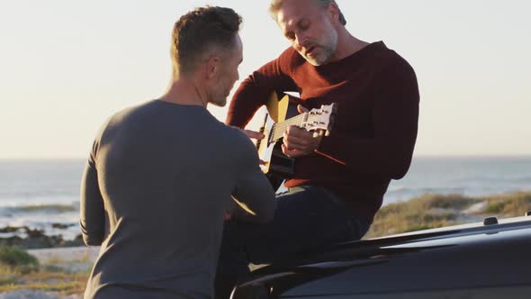 Happy caucasian gay male couple sitting on car playing guitar and talking at the beach