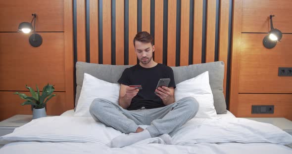 Young Man Sitting on the Bed and Pays for Purchases Online Using a Smartphone Enters Credit Card