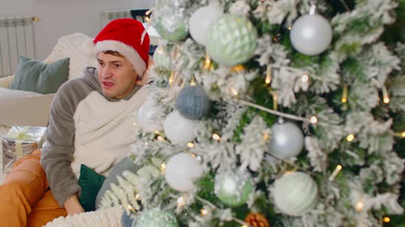 Sad Man in Santa Hat Sitting on Couch Near Christmas Tree in Living Room
