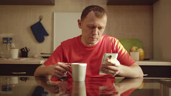 The Guy Reads the Message in His Mobile Phone While Sitting in the Kitchen Drinking Tea