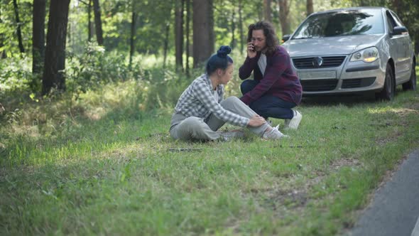 Wide Shot Injured Woman Sitting on Grass with Man Running From Car Calling Ambulance