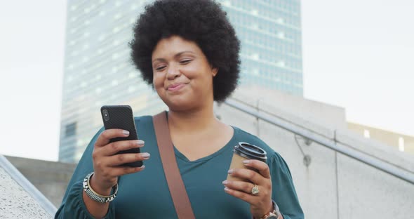 Happy plus size biracial woman using smartphone and drinking coffee