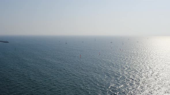 Multiple windsurfers with red sails surf the clear Mediterranean sea reflecting sunlight from the br