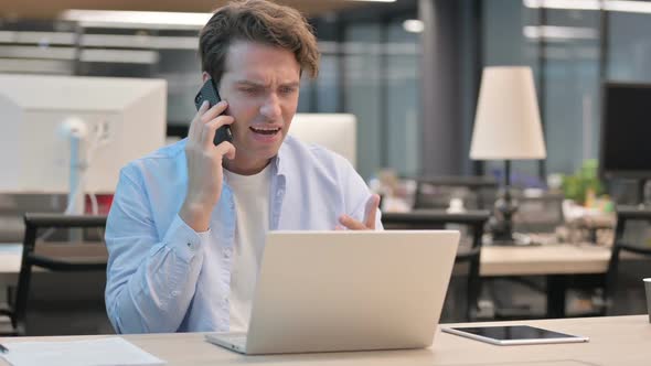 Angry Man Talking on Smartphone While Using Laptop in Office