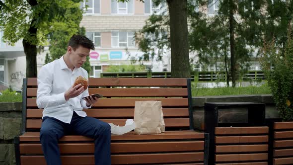 Hungry Young Man in Suit Gets a Hamburger and Eats It When Talks on Phone