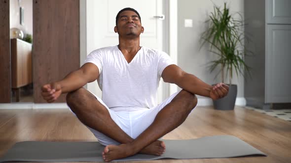 Relaxed AfricanAmerican Man with Closed Eyes Meditating at Home Sitting in Lotus Position on Mat