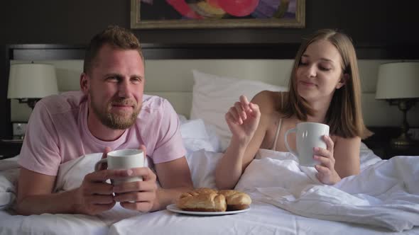 Man and Woman Are Eating Croissants with Coffee Lying in Bed in Morning