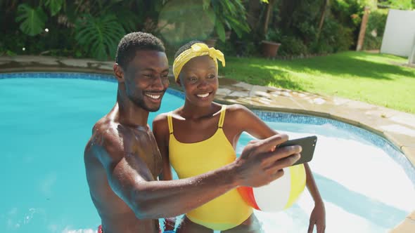 Happy african american couple standing in swimming pool taking selfie and smiling in sunny garden