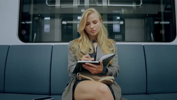 Young Blonde Girl Sitting in Metro Train Tired After Work Writing in Journal