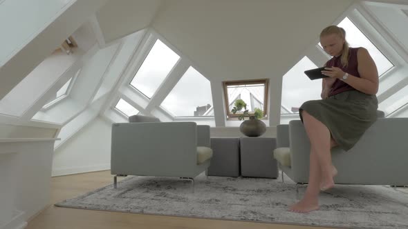 View of Young Blond Woman Sitting on the Side of Arm Chairs Using Tablet Inside of Room in a Cube