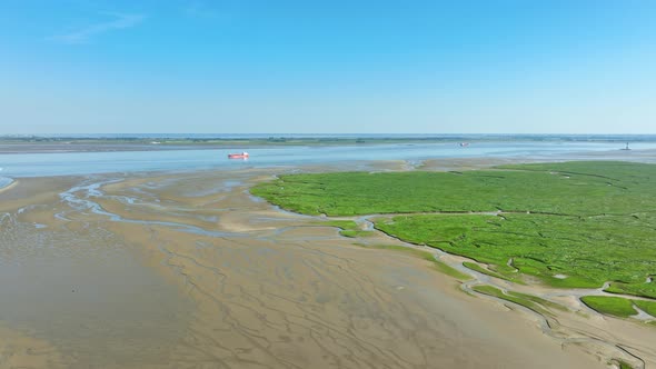 Aerial shot flying towards a red cargo ship passing by vibrant green wetlands along a river under a