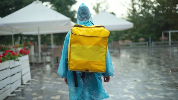 Back View Courier in Rain Coat and Coronavirus Face Mask with Backpack Walking on City Street