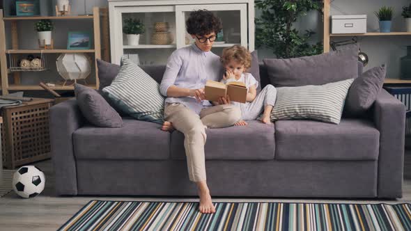 Young Woman Reading Book To Her Son Sitting on Sofa in Studio Apartment Together