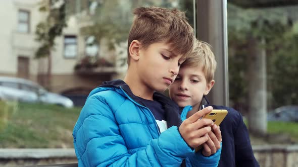 Two Boys Are Sitting At The Bus Stop. Playing Smartphones.