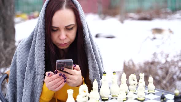 Young Woman Playing Chess and Browsing Smartphone in Yard