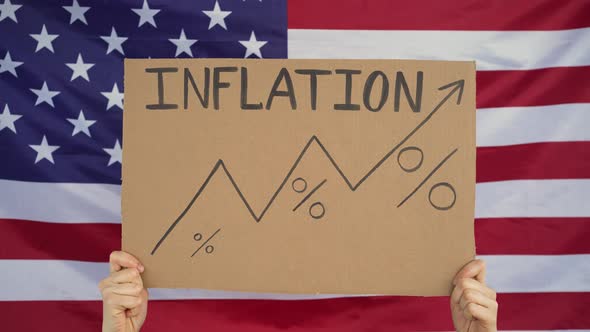 Hands Raise a Sign That Says Inflation and Graph Upward Against the Background of the American Flag