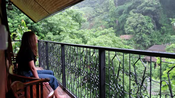 A woman sitting on balcony and looking at the rain over the tree