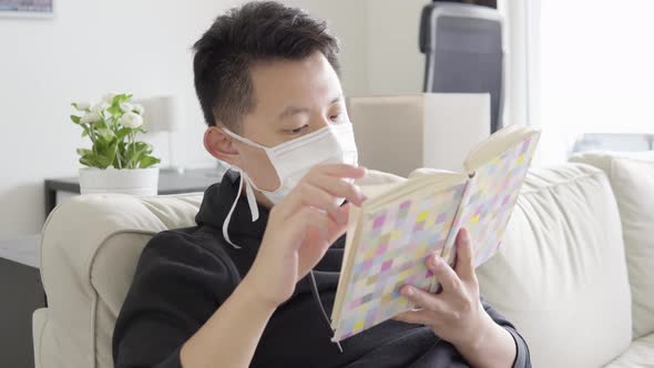 A Young Asian Man in a Face Mask Reads a Book As He Sits on a Couch at Home - Closeup