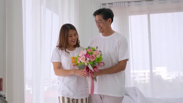 Asian man surprising his lover by giving a flower bouquet in bedroom.