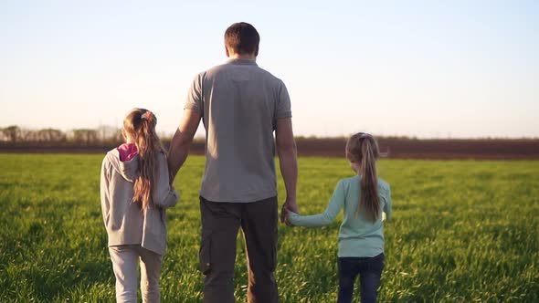 Backside View of a Strong Young Father Walking By a Green Meadow with Two Little Daughters
