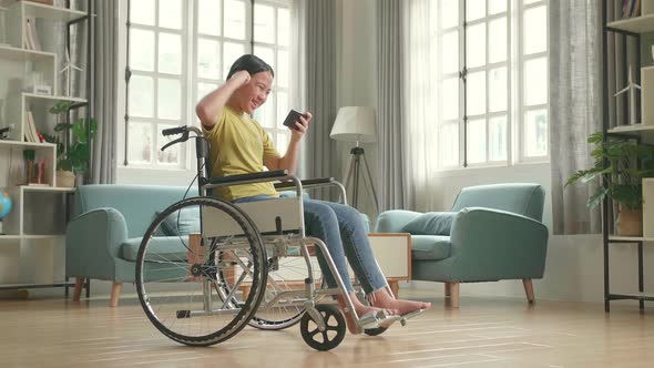 Excited Asian Kid Girl Sitting On Wheelchair Playing Video Game On Mobile Phone And Celebrating