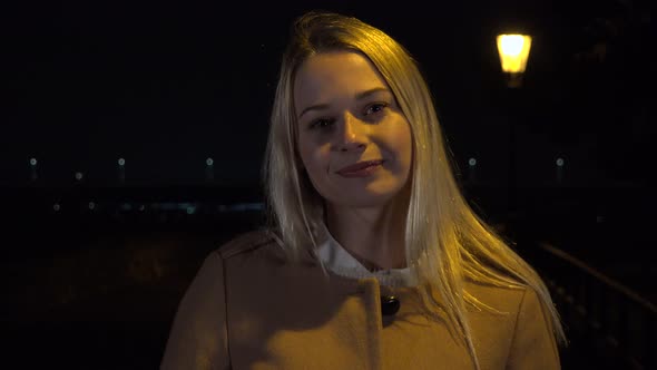 A Young Beautiful Woman Waves at the Camera with a Smile in an Urban Area at Night