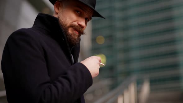 a Mustachioed and Bearded Stylish Man in a Hat and a Dark Coat Holds a Cigarette Looks at the Camera