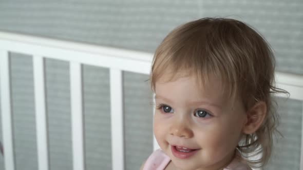 Close Up of Happy Little Girl with Curly Hair Stands and Smiles in White Crib