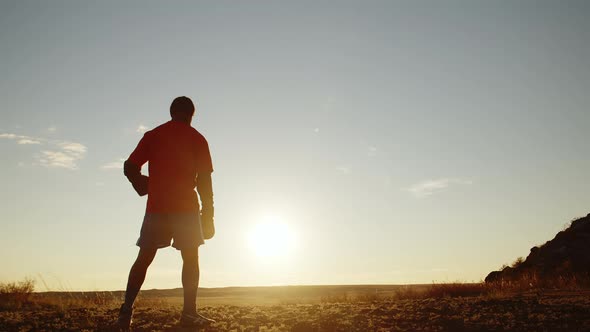 Male Athlete Stands in Sportswear and Boxing Gloves on Hill Against Background Sunset Sky Raises One