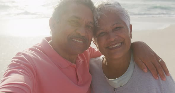 Happy hispanic senior couple embracing, taking selfie on beach at sunset
