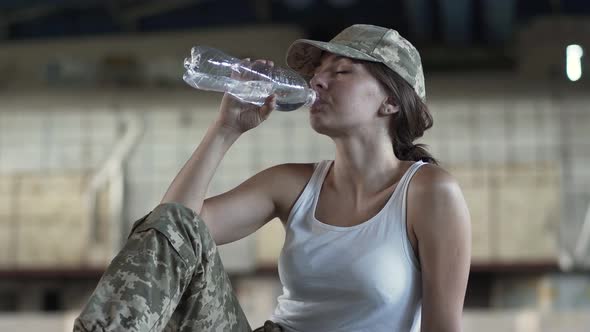 Cute Young Woman in Military Uniform Drinking Water From the Bottle Sitting on the Floor in Dusty