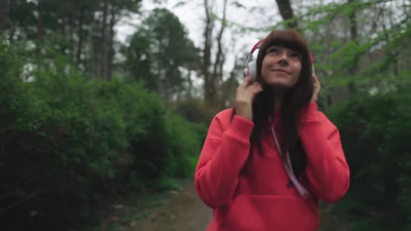 A young, cheerful woman with headphones on listens to music while walking in the park.