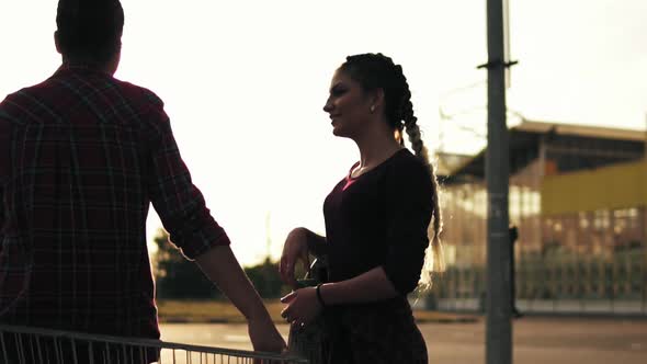 Side View of a Sexy Girl with Long Braids Holding a Skateboard and Talking to Her Girlfriend That is