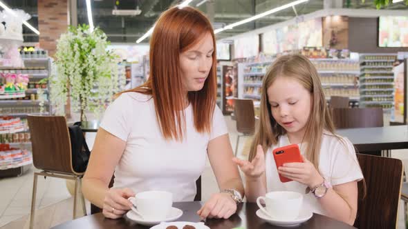 Girl and Woman Mother Sitting in Restaurant, Touching and Watching Smartphone Together