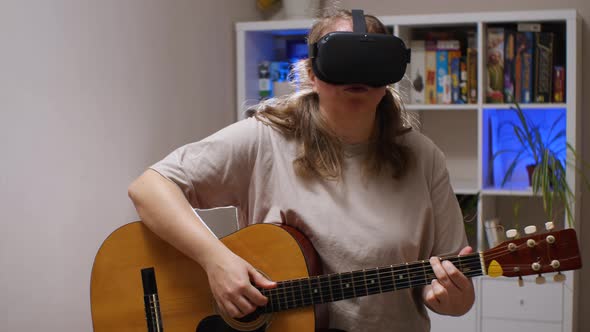A girl wearing VR glasses plays an acoustic guitar while sitting in a room