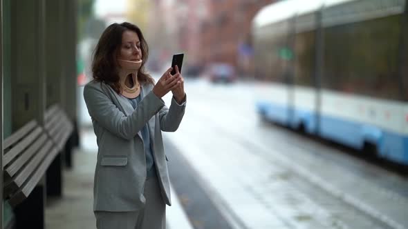 A Middle-aged Woman in a Jacket Stands By a City Road During the Day, Looking at Her Mobile Phone